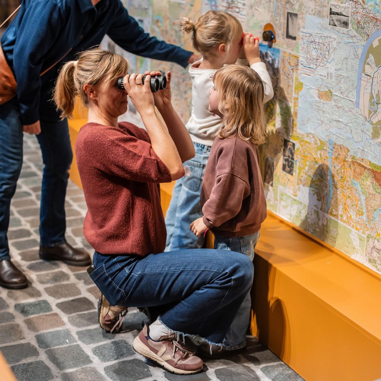 Mama kijkt door een verrekijker, twee kindjes kijken naar landkaarten